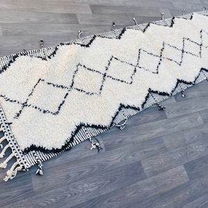 May include: A rectangular, off-white rug with a geometric black zigzag pattern. The rug has a shaggy texture and is trimmed with black and white tassels on the ends. The rug is placed on a grey wooden floor.