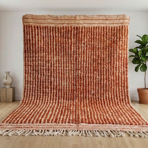 Puede incluir: Una alfombra rectangular grande con rayas verticales en tonos óxido y crema. La alfombra tiene un borde con flecos y se exhibe contra una pared blanca. La alfombra está hecha de un material tejido y es adecuada para una sala de estar o un dormitorio.