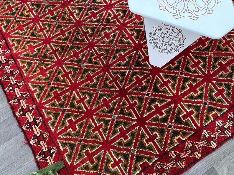 K&ouml;nnte beinhalten: Ein roter, gr&uuml;ner und cremefarbener Teppich mit geometrischem Muster. Der Teppich hat einen Fransenrand.