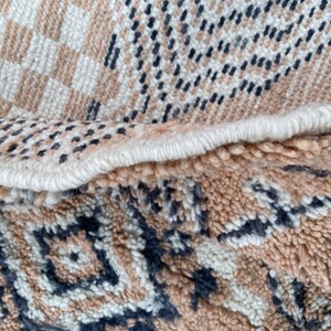 May include: Close-up of a woven rug with a geometric pattern. The rug features a combination of beige, white, and dark blue colors. The design includes checkered and diamond shapes, with a textured, looped pile.