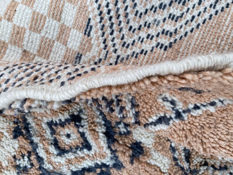 May include: Close-up of a woven rug with a geometric pattern. The rug features a combination of beige, white, and dark blue colors. The design includes checkered and diamond shapes, with a textured, looped pile.