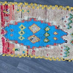 Puede incluir: Una alfombra marroqu&iacute; colorida con un patr&oacute;n geom&eacute;trico azul, amarillo, rojo y verde. La alfombra tiene un fondo blanco y est&aacute; hecha de lana.