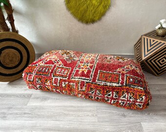 May include: A red, orange, and white patterned floor cushion with a fluffy texture. The cushion is rectangular and has a traditional Moroccan design.