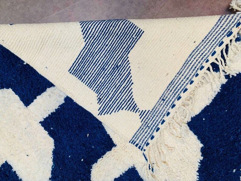 May include: A white and blue woven rug with a geometric pattern. The rug has a fringe border.