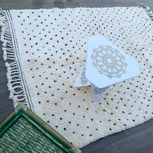 May include: White rug with black dots and a white triangular stool with a geometric design. The stool is sitting on the rug.
