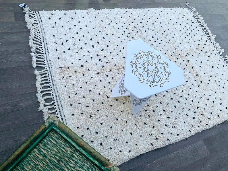 May include: White rug with black dots and a white triangular stool with a geometric design. The stool is sitting on the rug.