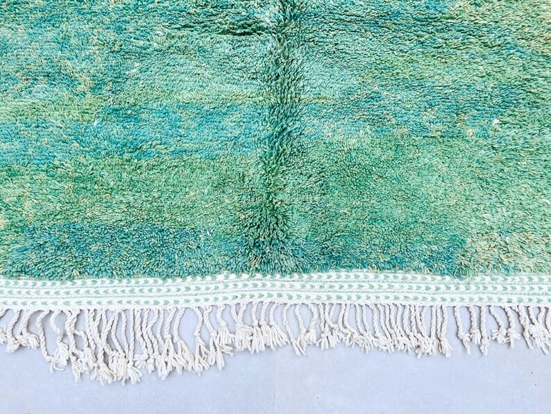 K&ouml;nnte beinhalten: Ein handgewebter Teppich in Teal- und Gr&uuml;nt&ouml;nen mit einem wei&szlig;en Fransenrand. Der Teppich hat einen subtilen Ombre-Effekt, der von einem dunkleren Teal zu einem helleren Gr&uuml;n &uuml;bergeht.