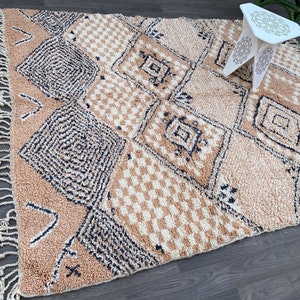 May include: A beige and cream-colored Moroccan rug with geometric diamond and square patterns. The rug has a textured appearance with fringe detailing along the edges. A small white decorative table with a patterned top is in the background.