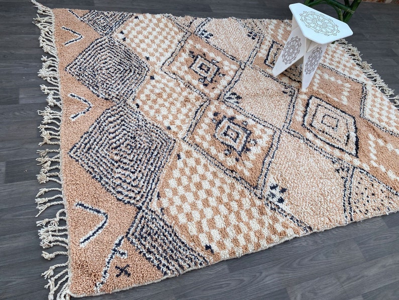 May include: A beige and cream-colored Moroccan rug with geometric diamond and square patterns. The rug has a textured appearance with fringe detailing along the edges. A small white decorative table with a patterned top is in the background.