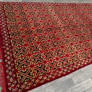 K&ouml;nnte beinhalten: Ein roter Teppich mit einem geometrischen Muster aus schwarzen und goldenen Kreuzen. Der Teppich hat einen Fransenrand.