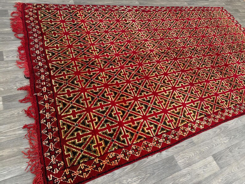K&ouml;nnte beinhalten: Ein roter Teppich mit einem geometrischen Muster aus schwarzen und goldenen Kreuzen. Der Teppich hat einen Fransenrand.
