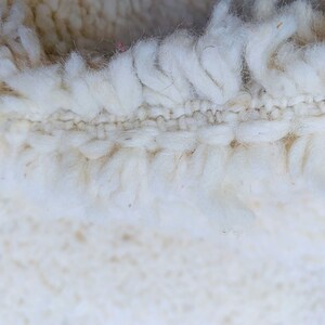Op de afbeelding: Close-up van een wit, pluche tapijt met een geweven rand. Het tapijt is gemaakt van zachte, natuurlijke vezels.