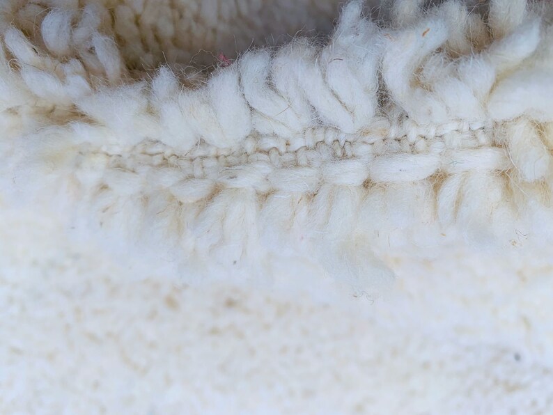 Op de afbeelding: Close-up van een wit, pluche tapijt met een geweven rand. Het tapijt is gemaakt van zachte, natuurlijke vezels.