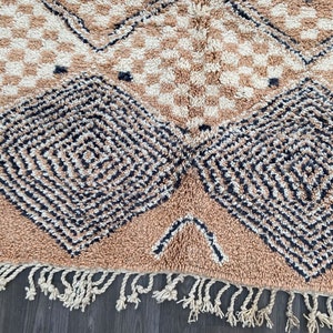 May include: Close-up of a beige and cream-colored rug with a geometric pattern. The rug features diamond and square designs in black and cream, with a fringe along the bottom edge. The rug is made of a soft, textured material.