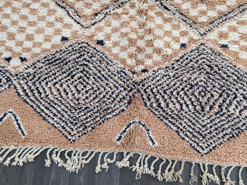 May include: Close-up of a beige and cream-colored rug with a geometric pattern. The rug features diamond and square designs in black and cream, with a fringe along the bottom edge. The rug is made of a soft, textured material.