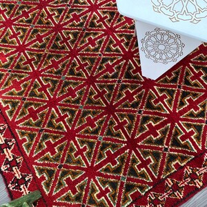 K&ouml;nnte beinhalten: Ein roter, gr&uuml;ner und cremefarbener Teppich mit geometrischem Muster. Der Teppich hat einen Fransenrand.