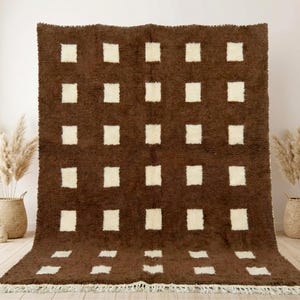 May include: A large, rectangular brown rug with a repeating pattern of cream-coloured squares. The rug has a shaggy texture and a fringe along the bottom edge. The rug is displayed indoors.