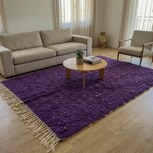 May include: A large, rectangular purple rug with a textured surface and cream-colored fringe. The rug is placed in a living room setting, with a beige sofa, wooden coffee table, and a light-colored armchair.