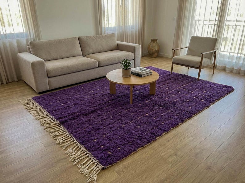 May include: A large, rectangular purple rug with a textured surface and cream-colored fringe. The rug is placed in a living room setting, with a beige sofa, wooden coffee table, and a light-colored armchair.