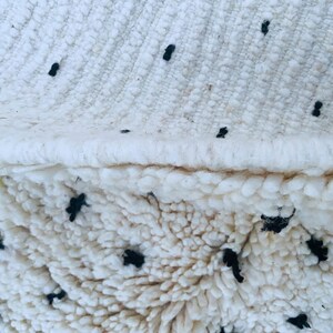 May include: Close-up of a white fluffy rug with black dots. The rug has a woven edge.