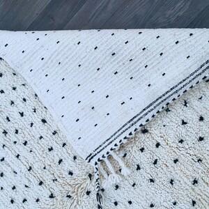 May include: A white and black Moroccan rug with a fringe border. The rug has a textured surface with small black dots.