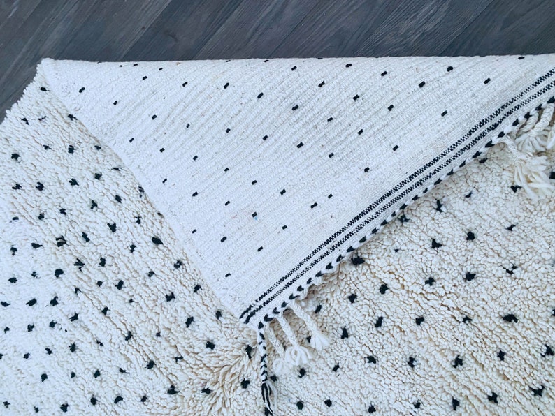 May include: A white and black Moroccan rug with a fringe border. The rug has a textured surface with small black dots.