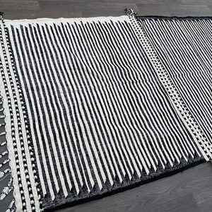 Striped Moroccan rug handwoven with black and white wool, creating clean vertical lines across the surface (see photos). Its minimalist pattern is enriched by fringed details, reflecting authentic Berber heritage. This durable carpet fits effortlessly into boho living rooms, modern apartments, or cozy bedrooms, adding warmth, structure, and cultural depth to your space.