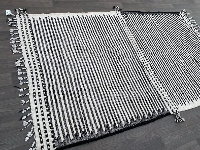 Striped Moroccan rug handwoven with black and white wool, creating clean vertical lines across the surface (see photos). Its minimalist pattern is enriched by fringed details, reflecting authentic Berber heritage. This durable carpet fits effortlessly into boho living rooms, modern apartments, or cozy bedrooms, adding warmth, structure, and cultural depth to your space.