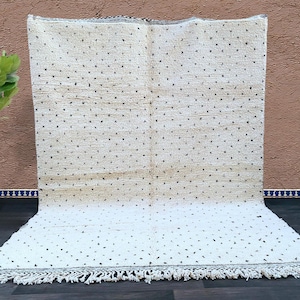 Op de afbeelding: Crèmekleurig tapijt met een stippenpatroon en gefranjerde randen. Het tapijt heeft een textuur en een gradiënteffect, van een beige bovenkant naar een witte onderkant. Geschikt voor interieurdecoratie.
