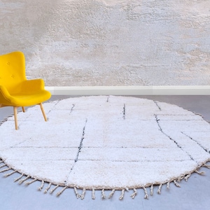 May include: A white round rug with a fringe border and a geometric pattern. The rug is on a gray floor in front of a textured wall. A yellow armchair is sitting on the rug.