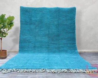 May include: A teal blue shag rug with a fringe border. The rug is laid out on a light gray floor.