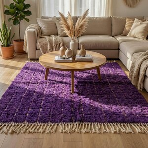 May include: A plush, rectangular purple rug with a textured, grid-like pattern. The rug has a cream-colored fringe along the bottom edge. A round wooden coffee table sits on the rug, with a beige sectional sofa in the background.