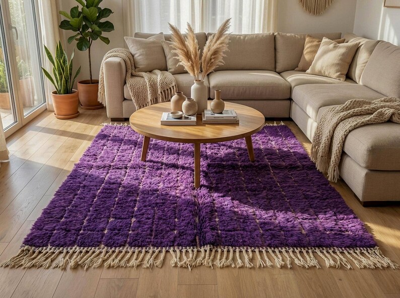 May include: A plush, rectangular purple rug with a textured, grid-like pattern. The rug has a cream-colored fringe along the bottom edge. A round wooden coffee table sits on the rug, with a beige sectional sofa in the background.