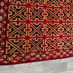 K&ouml;nnte beinhalten: Ein roter Teppich mit einem geometrischen Muster in Schwarz, Gr&uuml;n und Gelb. Der Teppich hat einen Fransenrand.
