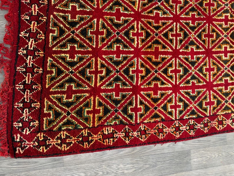 K&ouml;nnte beinhalten: Ein roter Teppich mit einem geometrischen Muster in Schwarz, Gr&uuml;n und Gelb. Der Teppich hat einen Fransenrand.