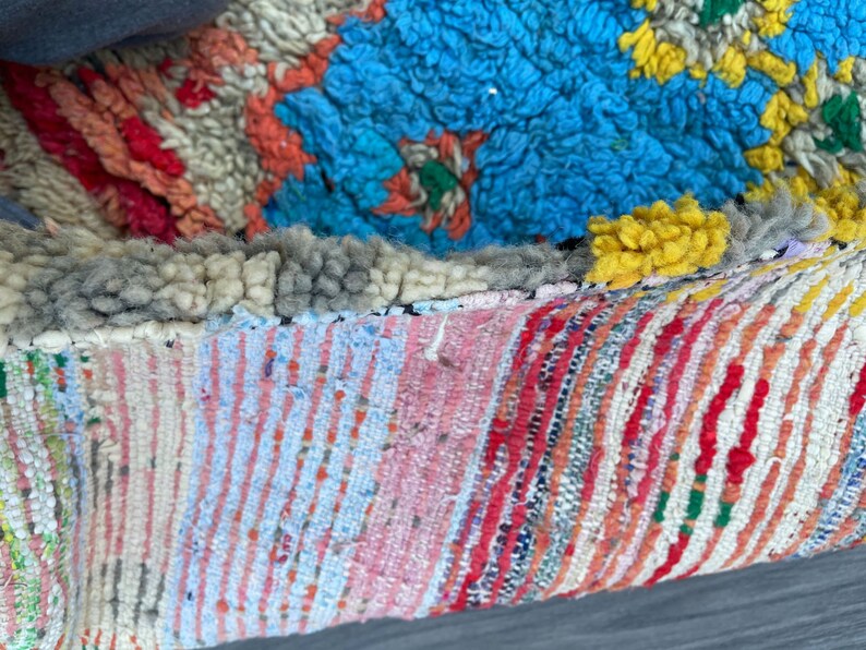 Puede incluir: Primer plano de una alfombra marroqu&iacute; colorida con una variedad de texturas y patrones. La alfombra est&aacute; hecha de lana y tiene un borde de flecos. La alfombra est&aacute; en tonos de azul, rojo, amarillo, blanco y gris.