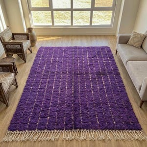 May include: A large, rectangular purple rug with a grid pattern of white lines. The rug has a shaggy texture and a fringe along the bottom edge. The rug is placed on a wooden floor in a room with natural light.