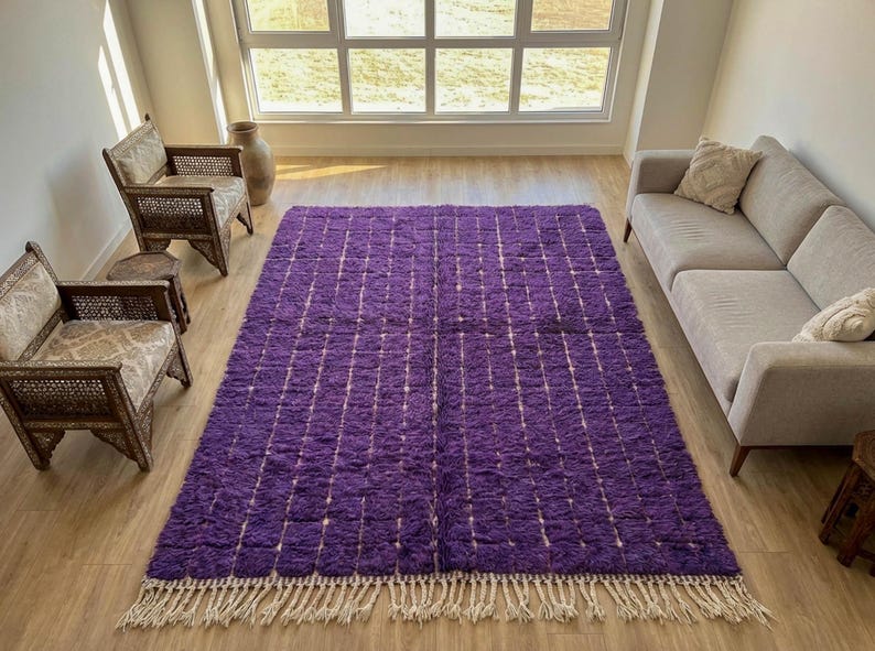 May include: A large, rectangular purple rug with a grid pattern of white lines. The rug has a shaggy texture and a fringe along the bottom edge. The rug is placed on a wooden floor in a room with natural light.