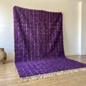 May include: A large, rectangular purple rug with a grid pattern of off-white lines. The rug has a thick, textured pile and a fringe along the bottom edge. A decorative vase is visible in the background.