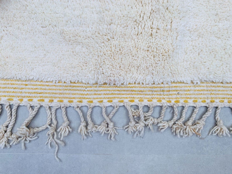 Op de afbeelding: Een witte badmat met een gele en witte gestreepte rand en een franje van witte kwastjes.