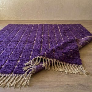 May include: A rectangular purple rug with a textured surface and a beige fringe. The rug has a pattern of vertical lines and is partially folded over, revealing a beige underside. The rug is on a wooden floor.