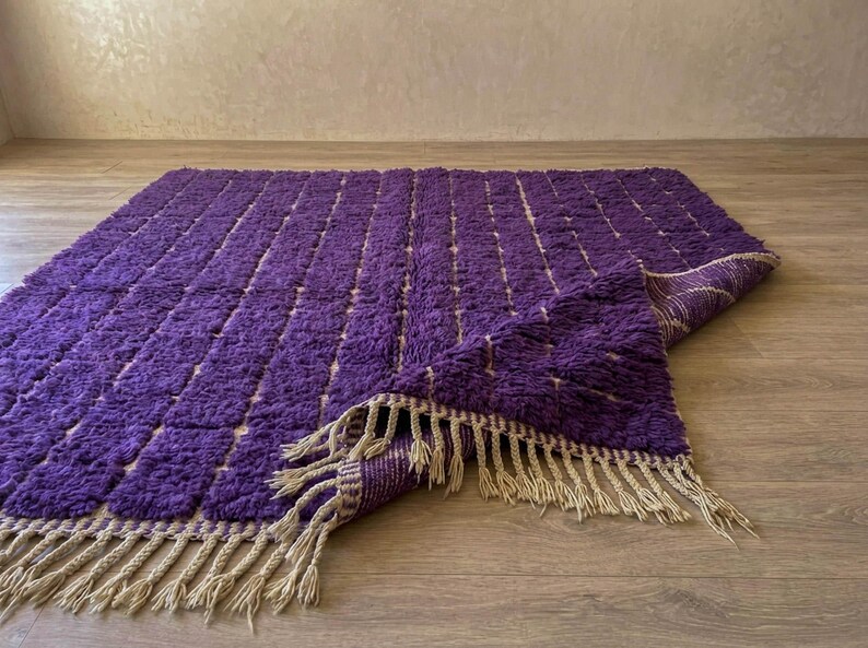 May include: A rectangular purple rug with a textured surface and a beige fringe. The rug has a pattern of vertical lines and is partially folded over, revealing a beige underside. The rug is on a wooden floor.