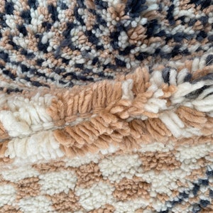 May include: Close-up of a textured rug with a mix of colors and patterns. The rug features a combination of beige, white, and navy blue hues. The design includes a checkerboard pattern and a speckled pattern, creating a visually interesting textile.