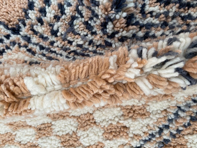 May include: Close-up of a textured rug with a mix of colors and patterns. The rug features a combination of beige, white, and navy blue hues. The design includes a checkerboard pattern and a speckled pattern, creating a visually interesting textile.