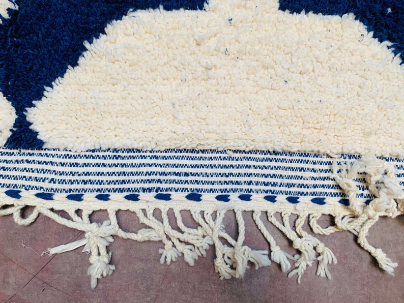 May include: A white and blue rug with a fringe border. The rug features a geometric pattern and is made from a soft, fluffy material.