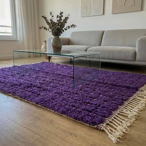 May include: A vibrant purple rug with a textured surface and a cream-colored fringe. The rug is placed on a wooden floor, with a glass coffee table and a gray sofa in the background. A vase with dried plants sits on the table.