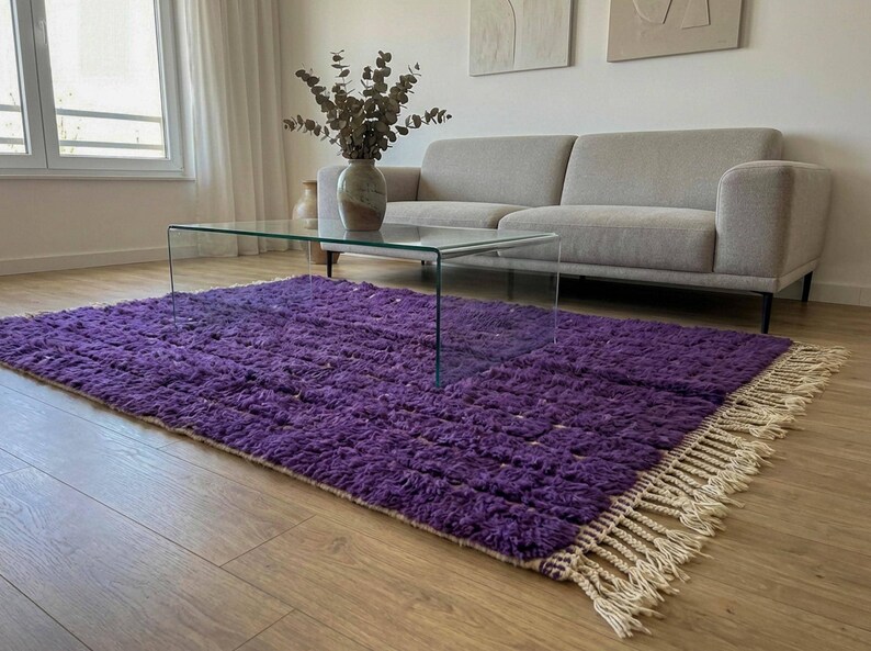 May include: A vibrant purple rug with a textured surface and a cream-colored fringe. The rug is placed on a wooden floor, with a glass coffee table and a gray sofa in the background. A vase with dried plants sits on the table.
