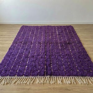 May include: A rectangular, plush purple rug with a grid pattern. The rug has a thick, textured pile and a beige fringe along the bottom edge. The rug is placed on a wooden floor.