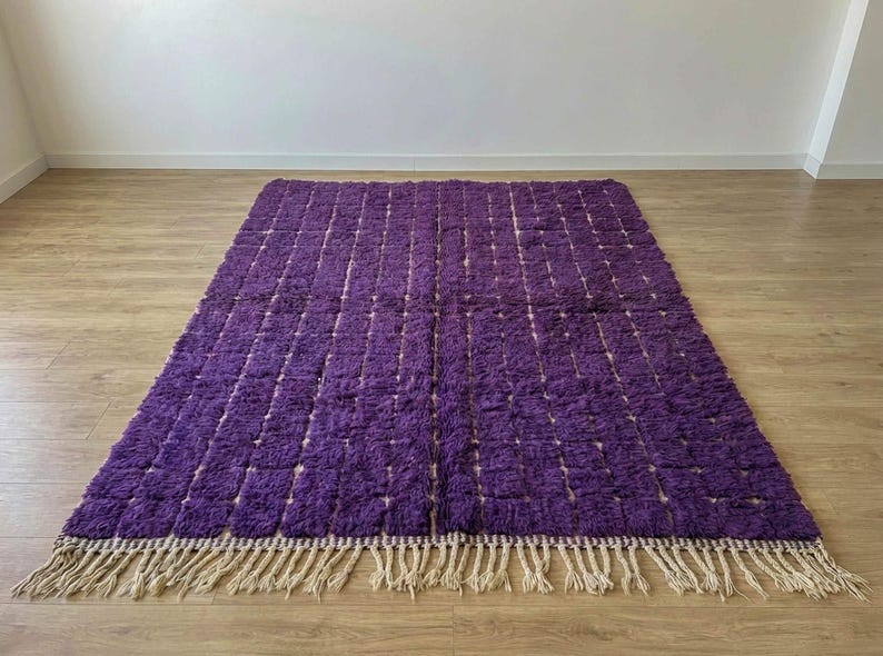 May include: A rectangular, plush purple rug with a grid pattern. The rug has a thick, textured pile and a beige fringe along the bottom edge. The rug is placed on a wooden floor.