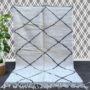 May include: A white Moroccan rug with a black diamond pattern. The rug has a fringe border and is made of wool.